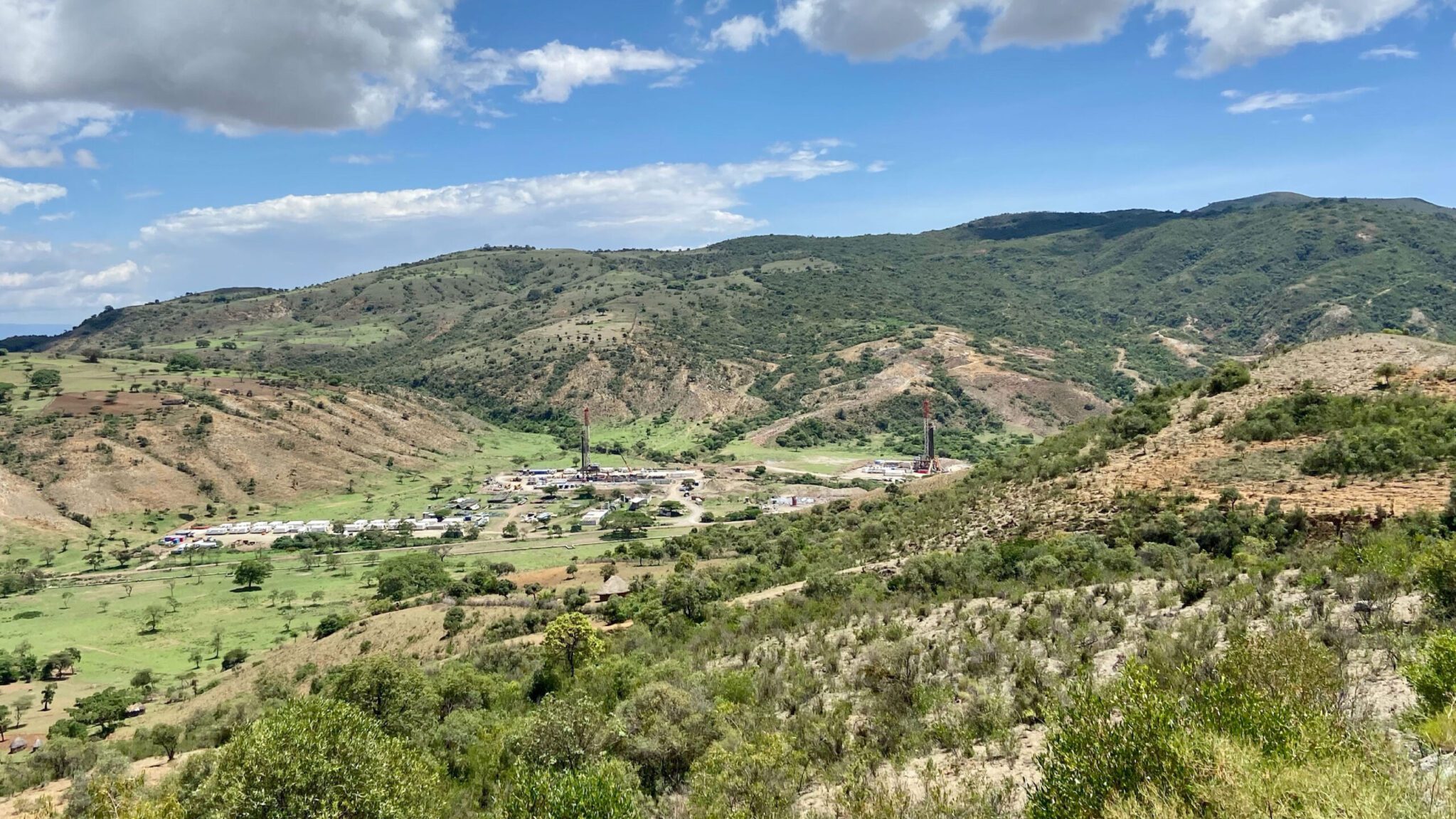Panoramic view of a small village nestled in a hilly green landscape, with scattered trees and mountains in the background under a partly cloudy sky