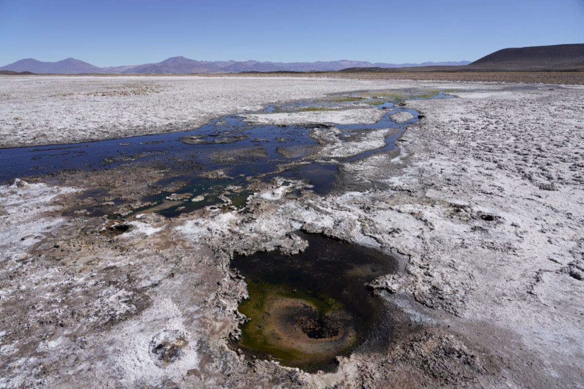 Arid salt flat landscape with shallow water pools and mineral deposits, surrounded by distant mountains under a clear sky