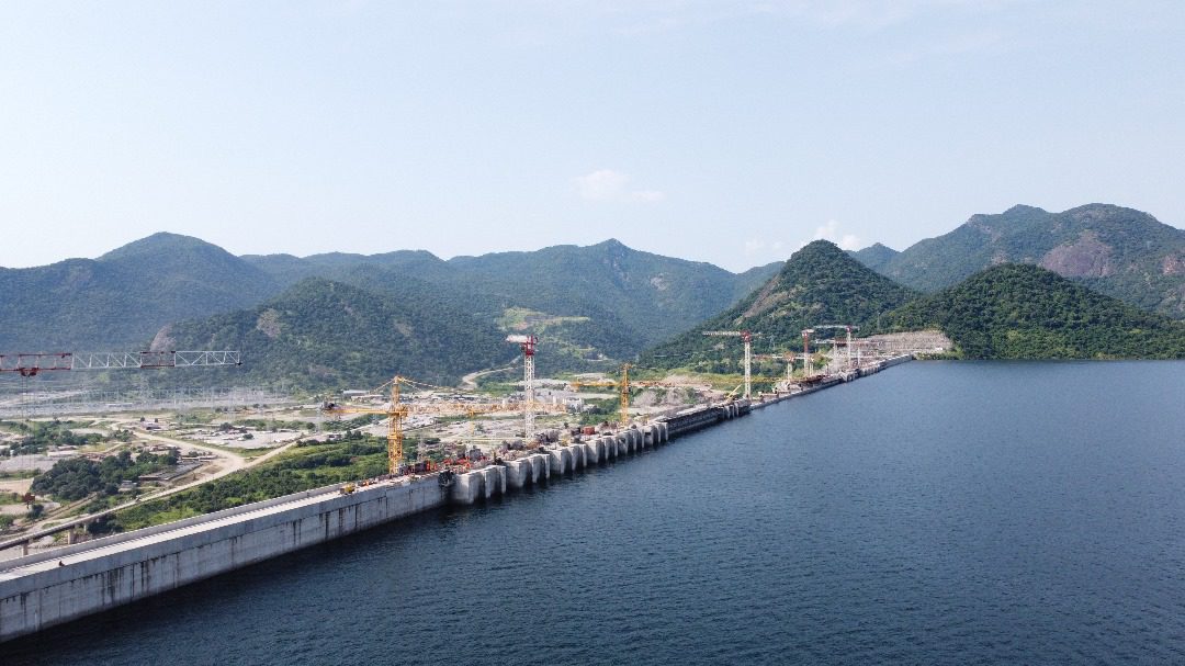Wide view of a large dam structure extending across a body of water, with cranes and construction activity visible, set against a backdrop of forested hills