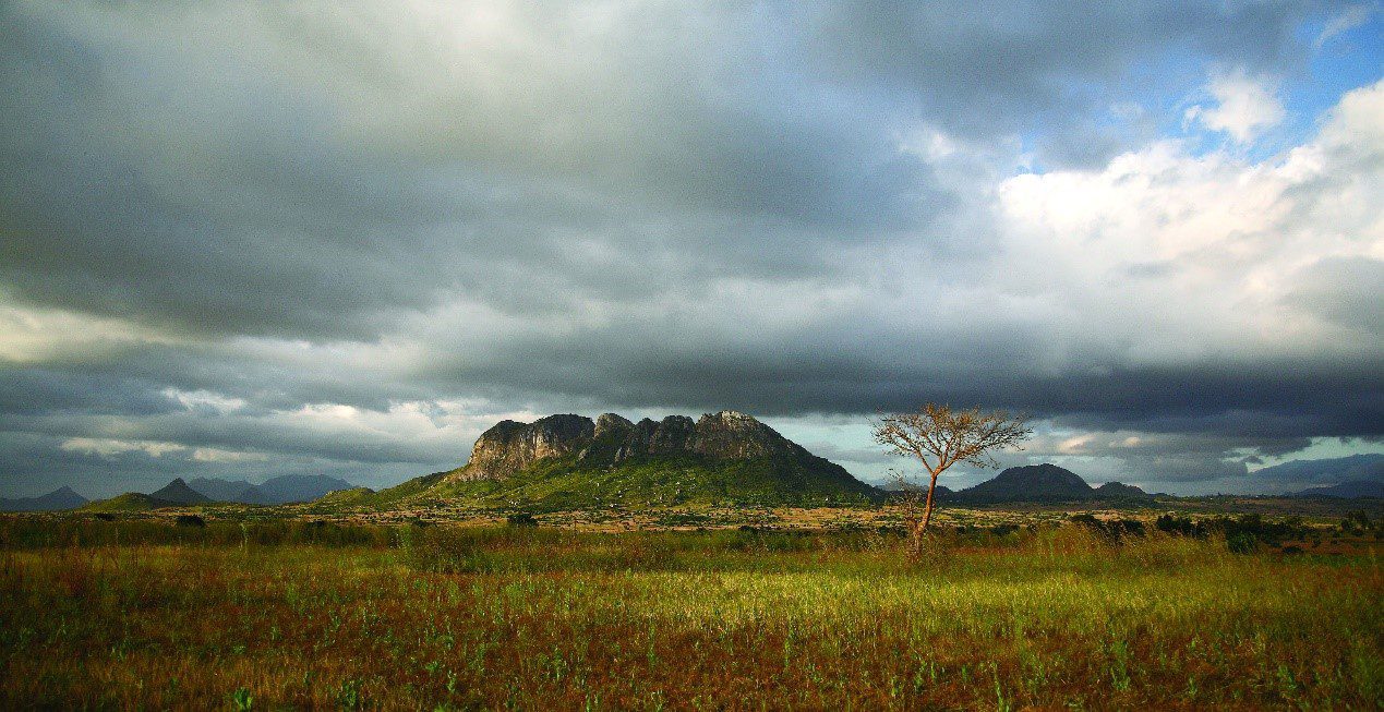 Isolated rocky hill covered with vegetation rising from a wide grassy plain, under a dramatic cloudy sky