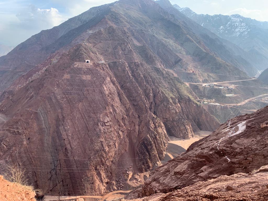 Mountain landscape with steep reddish-brown rocky slopes, a winding road, and snow-capped peaks in the background
