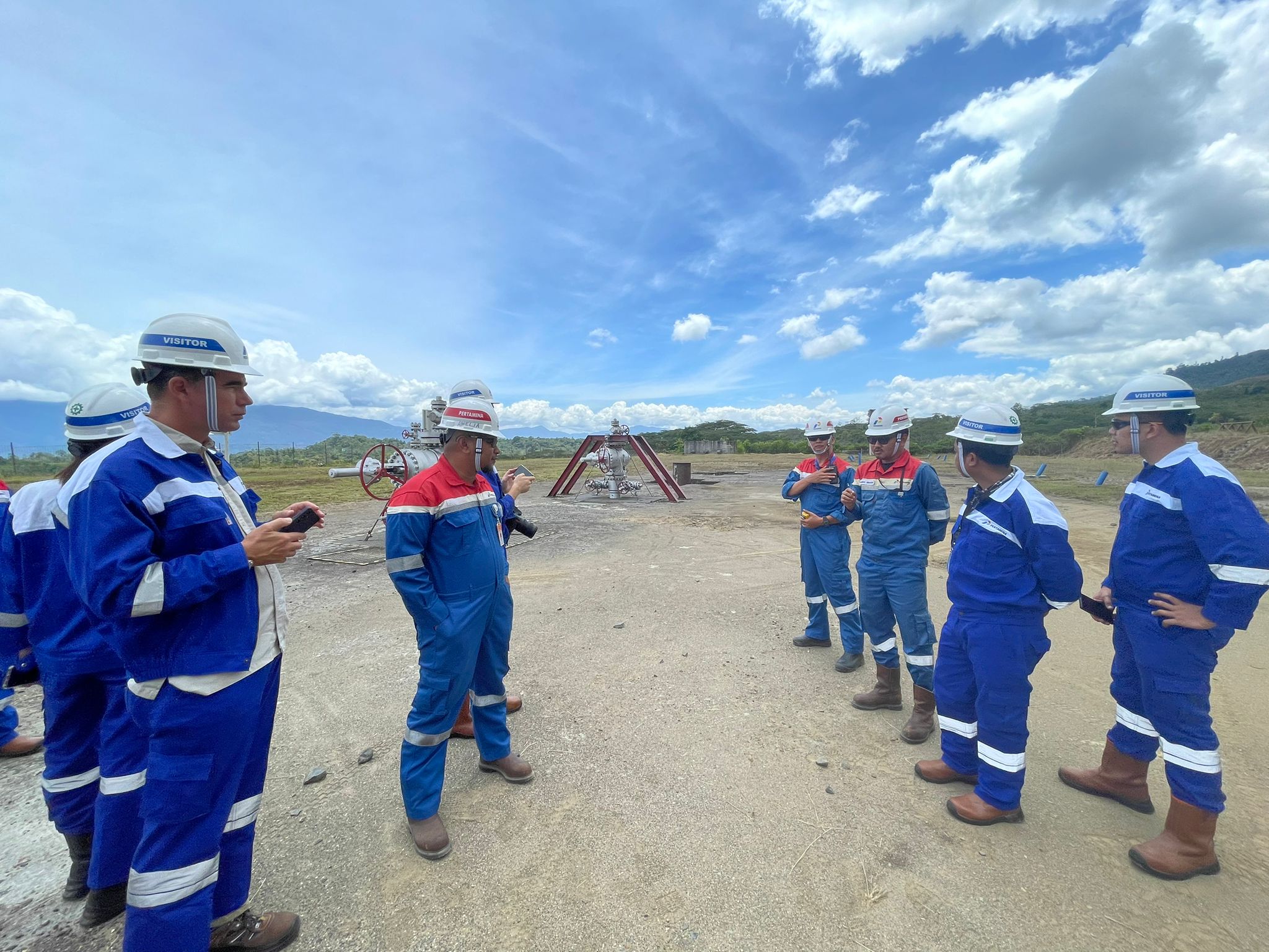 Group of workers in blue protective uniforms and helmets gathered outdoors at a construction or industrial site under a partly cloudy sky