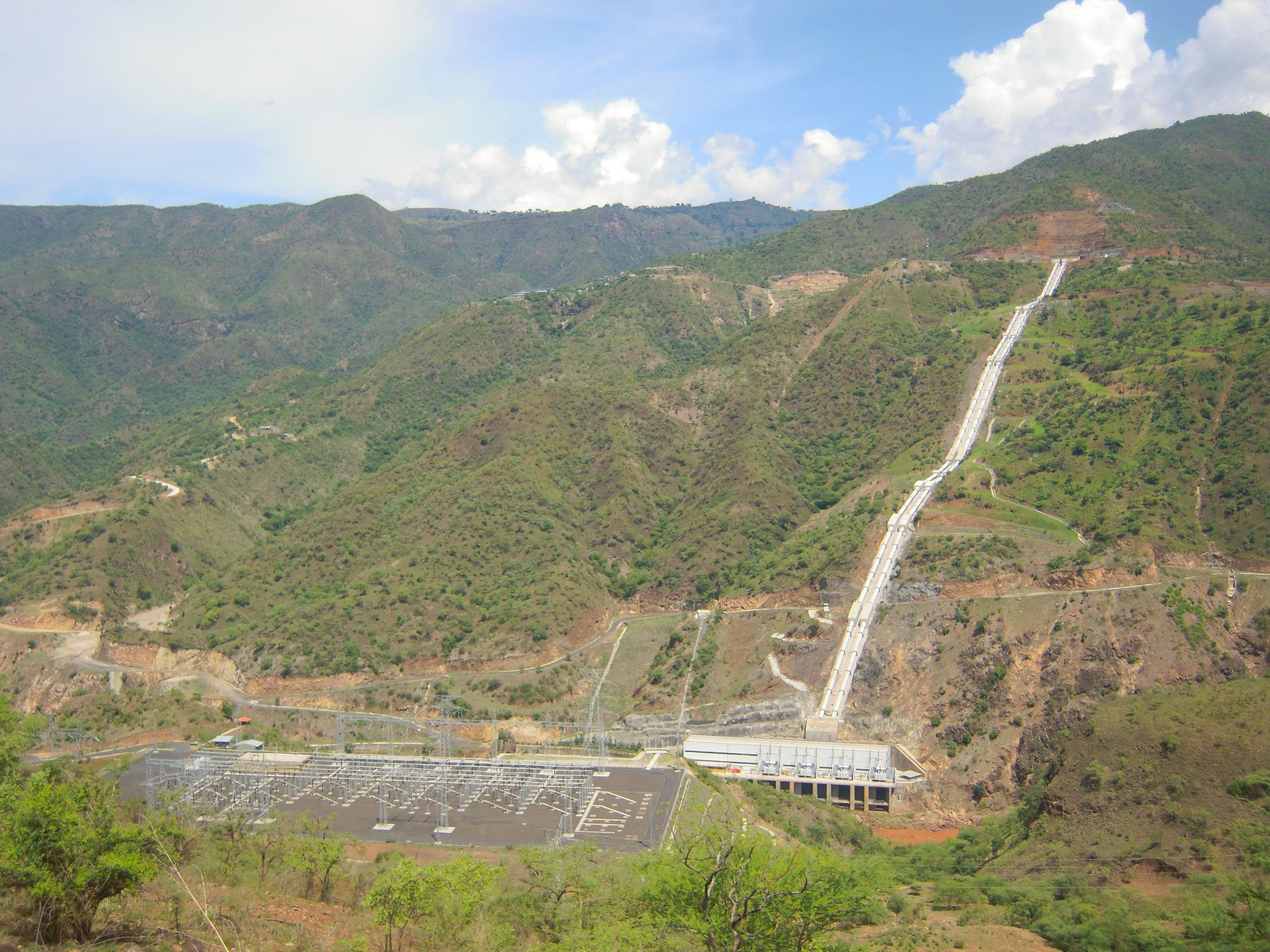 Panoramic view of a hydroelectric facility in a mountainous landscape, with a long penstock descending the hillside toward a power station