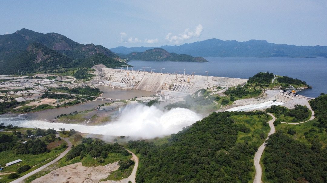 Aerial view of a massive dam structure with active water discharge, surrounded by lush hills and a large reservoir.