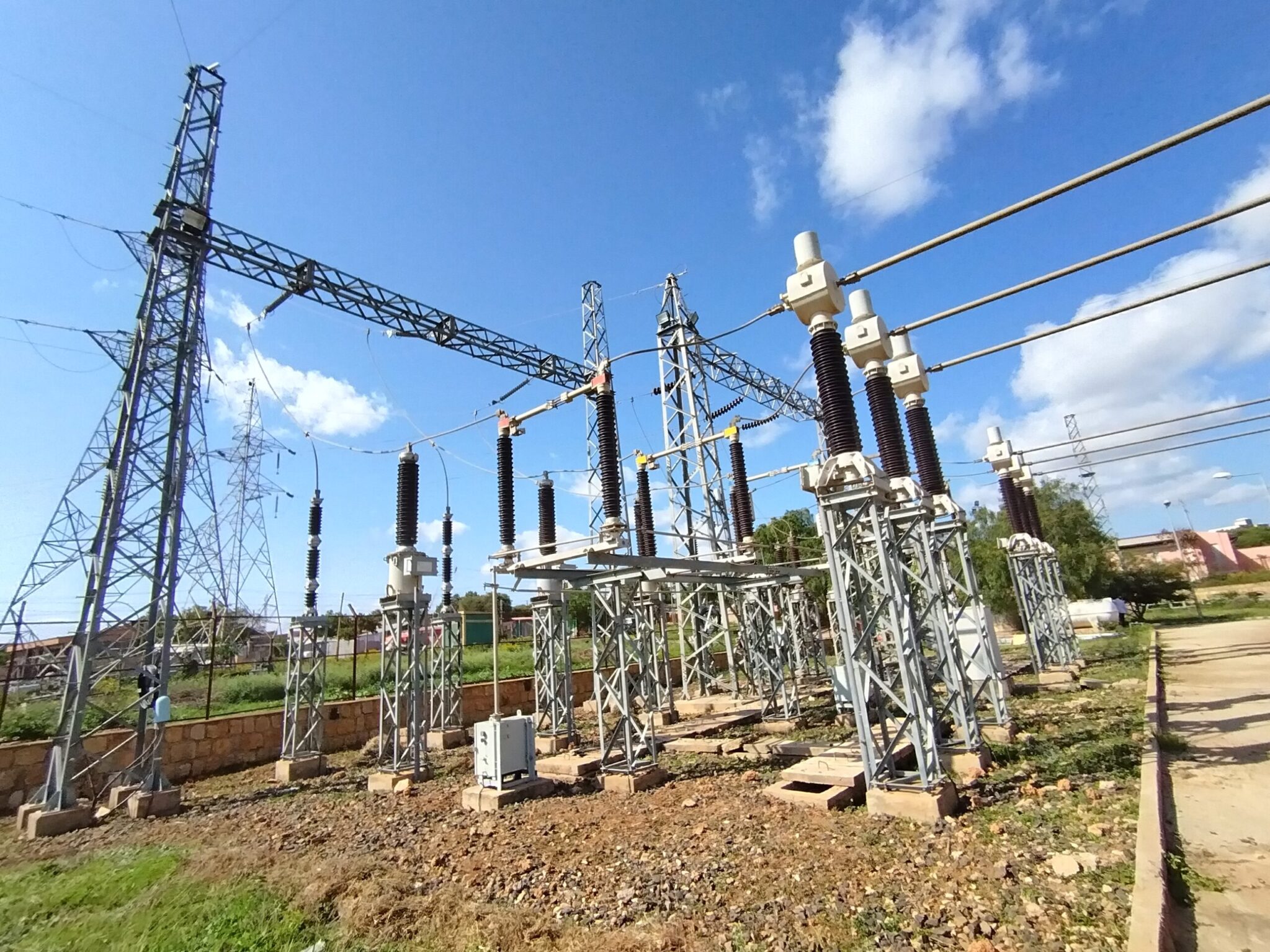 Outdoor electrical substation with high-voltage equipment, insulators, and transmission lines under a bright blue sky with scattered clouds