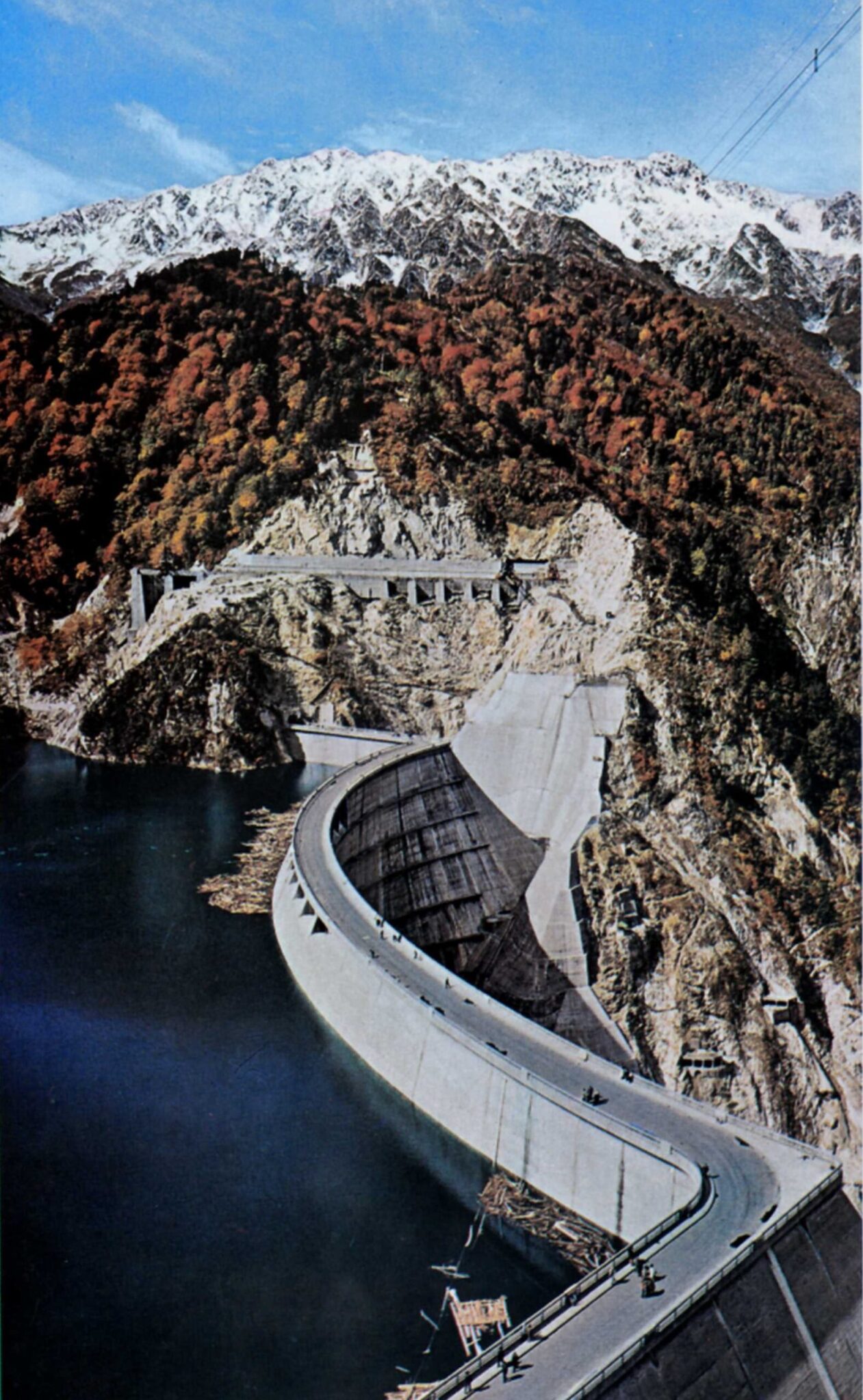 Aerial view of a large curved concrete dam in a mountainous landscape, surrounded by autumn-forest.
