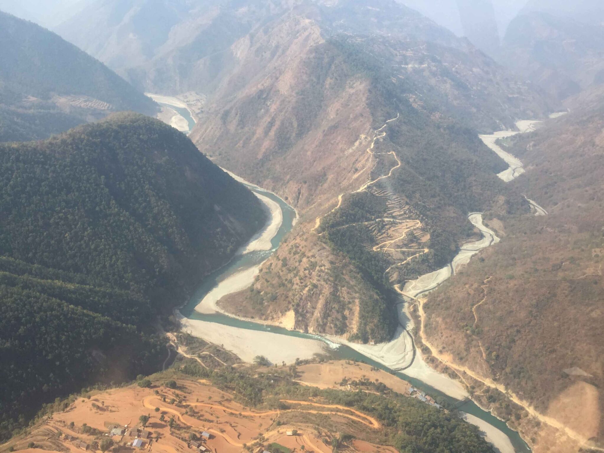 Aerial view of a river winding through steep mountainous terrain, with terraced farming visible on the slopes and construction works along the riverbank.