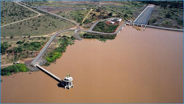 Aerial view of a dam and reservoir with brownish water, surrounded by arid land and infrastructure including a spillway and intake tower