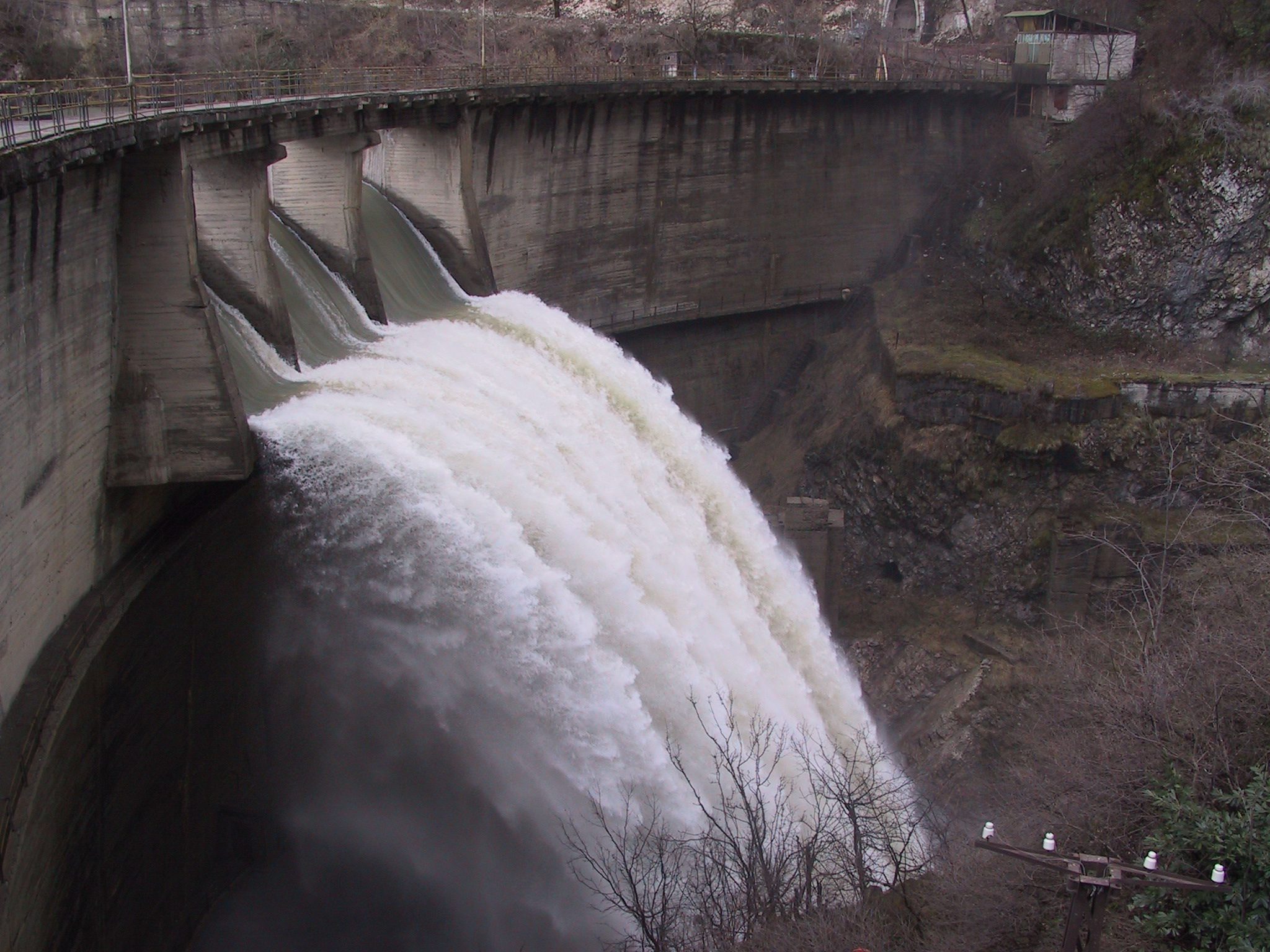 Powerful water discharge from a dam spillway, with water gushing through multiple gates into a rocky downstream area
