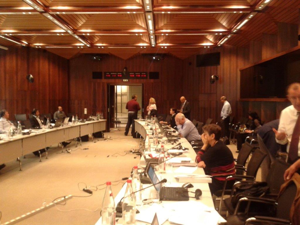 Formal meeting or conference in a large wood-paneled room, with participants seated at a U-shaped table, some standing or conversing during a break