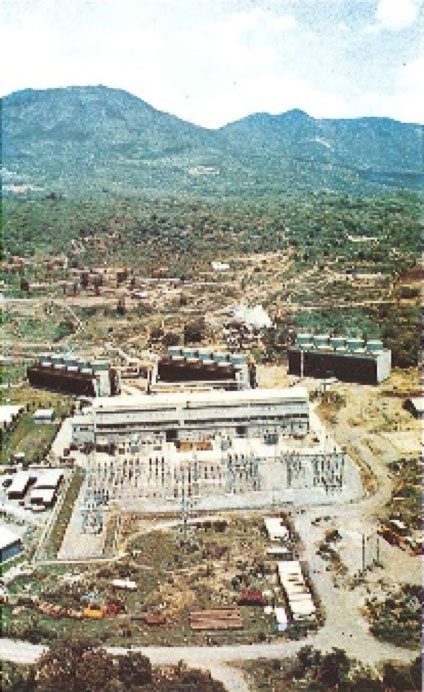 Aerial view of a geothermal power plant complex, located in a green valley surrounded by mountains.