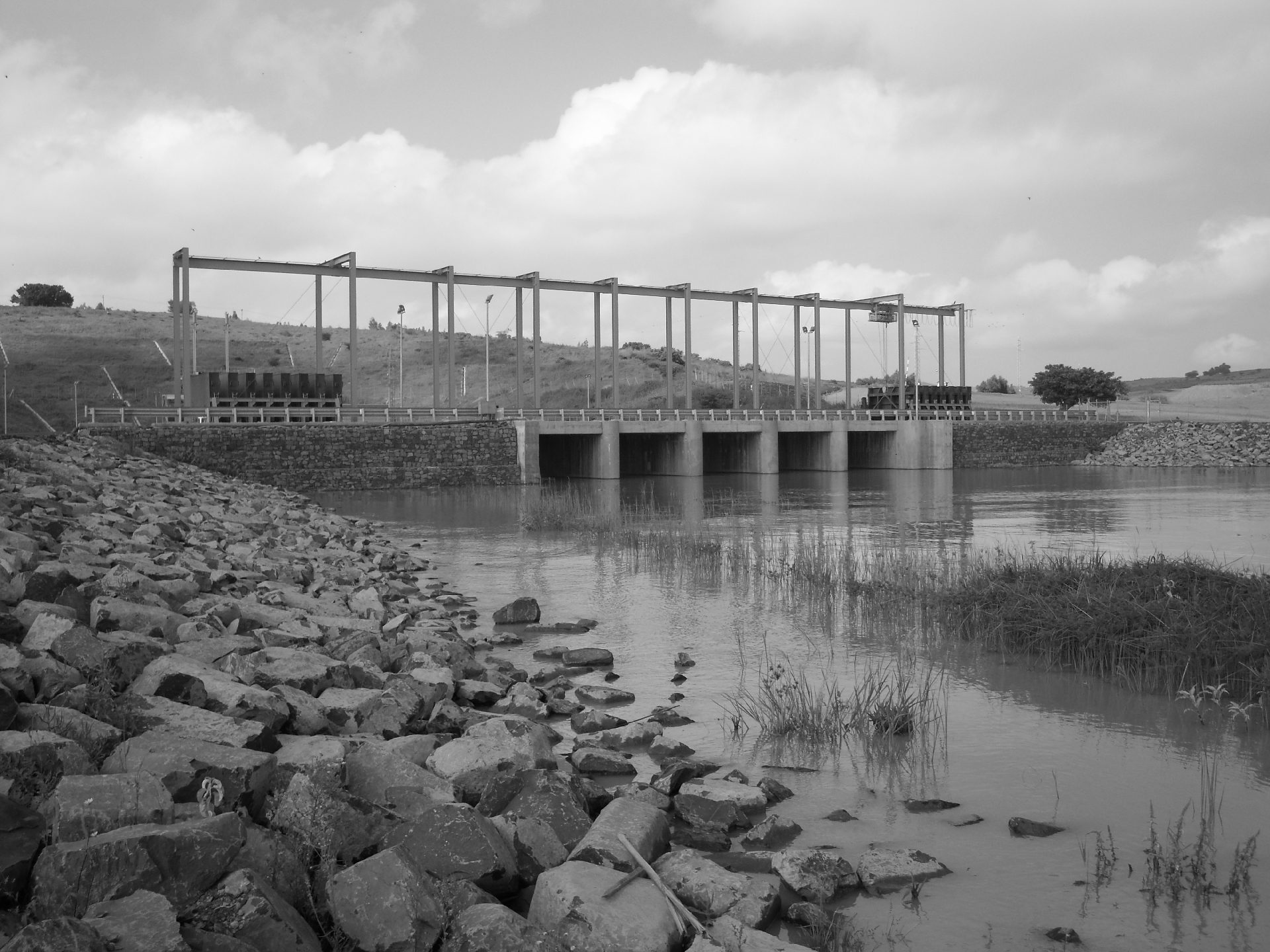 Black and white image of a small dam or water control structure with multiple gates, surrounded by rocky banks and calm water
