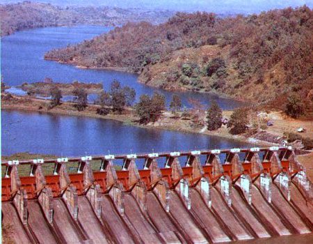 Aerial view of a large dam with multiple spillway gates, holding back a reservoir surrounded by forested hills