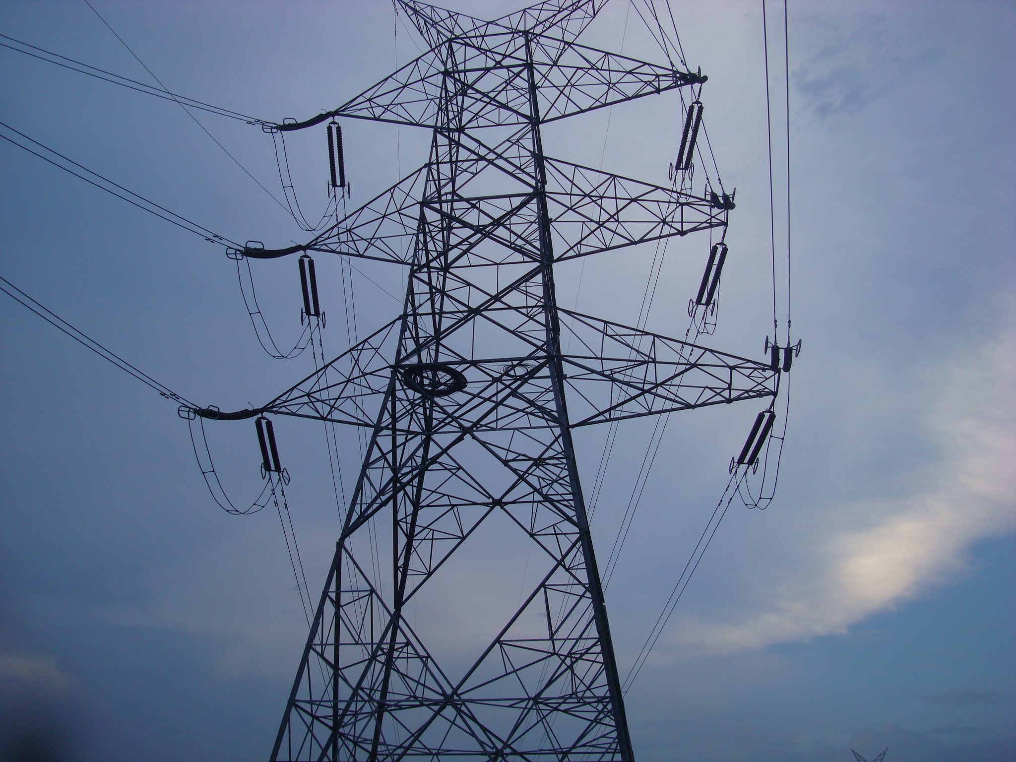 High-voltage transmission tower with multiple power lines, silhouetted against a cloudy evening sky