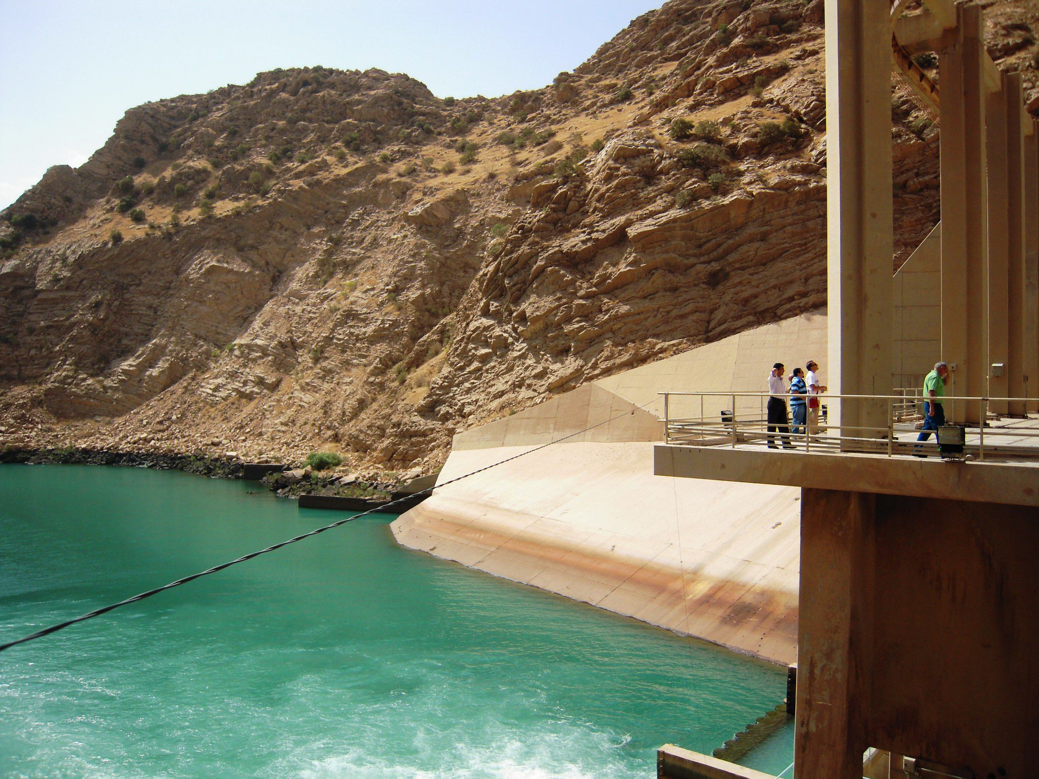 People standing on a viewing platform near a dam with turquoise water, set against a rocky, arid hillside