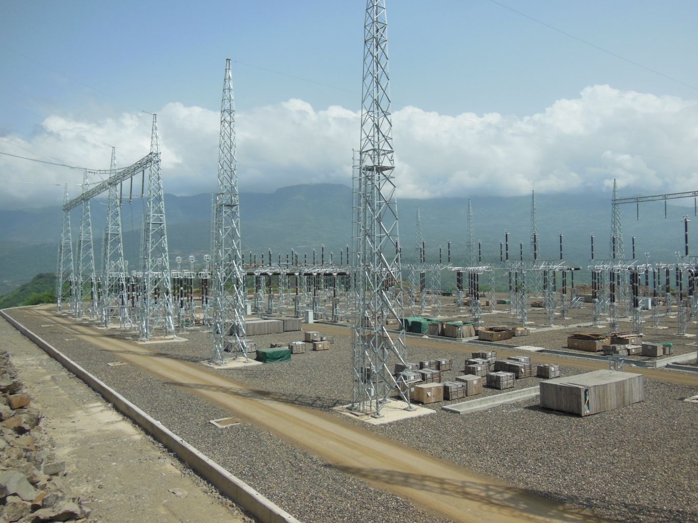 Wide view of an electrical substation under construction, with metal lattice towers, equipment foundations, and mountains in the background