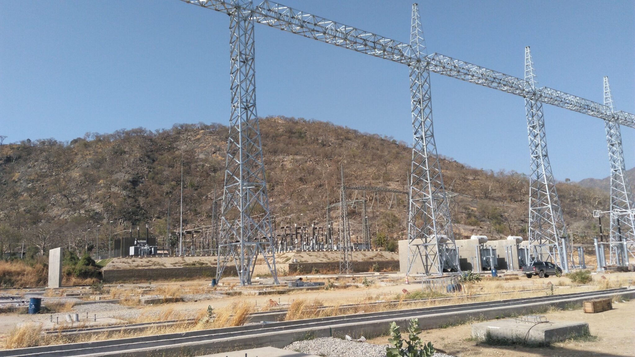 High-voltage electrical substation with metal lattice structures and transformers, located in a dry, hilly landscape under a clear sky