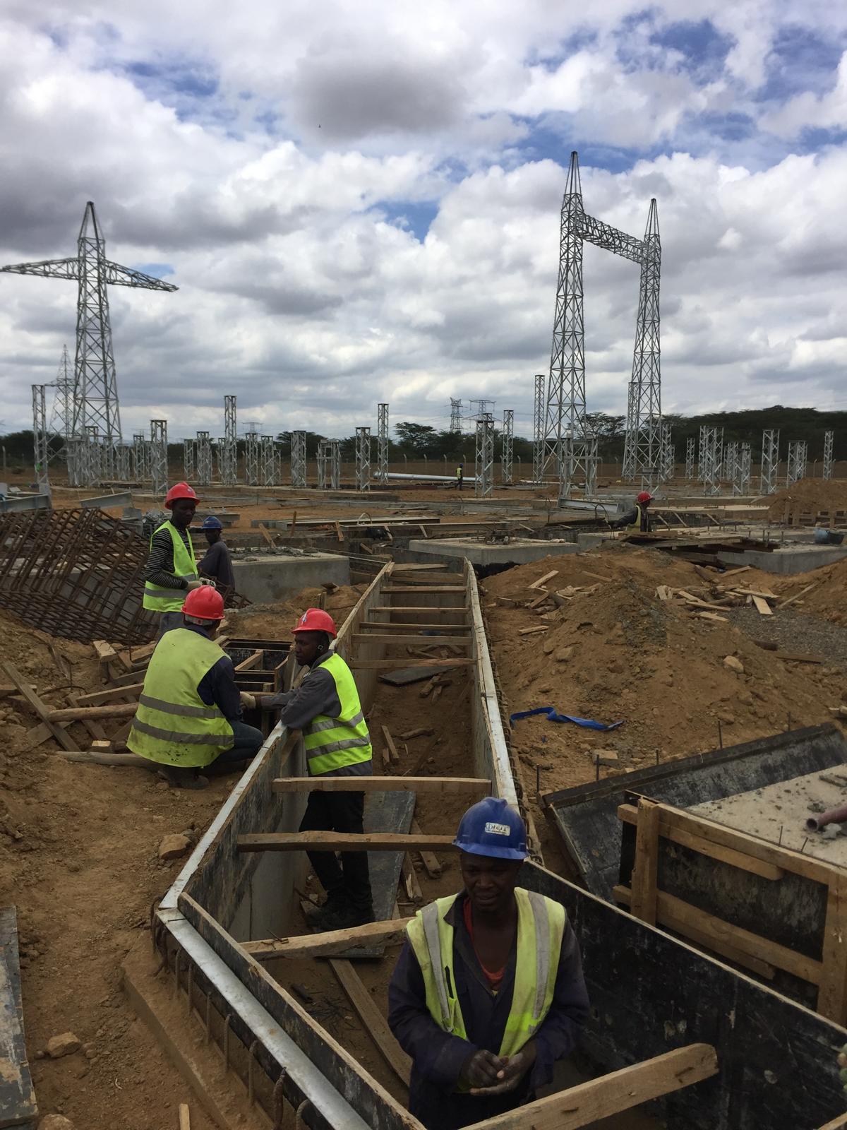 Construction workers wearing safety vests and helmets working on concrete foundations at an electrical substation site, with high-voltage transmission towers in the background