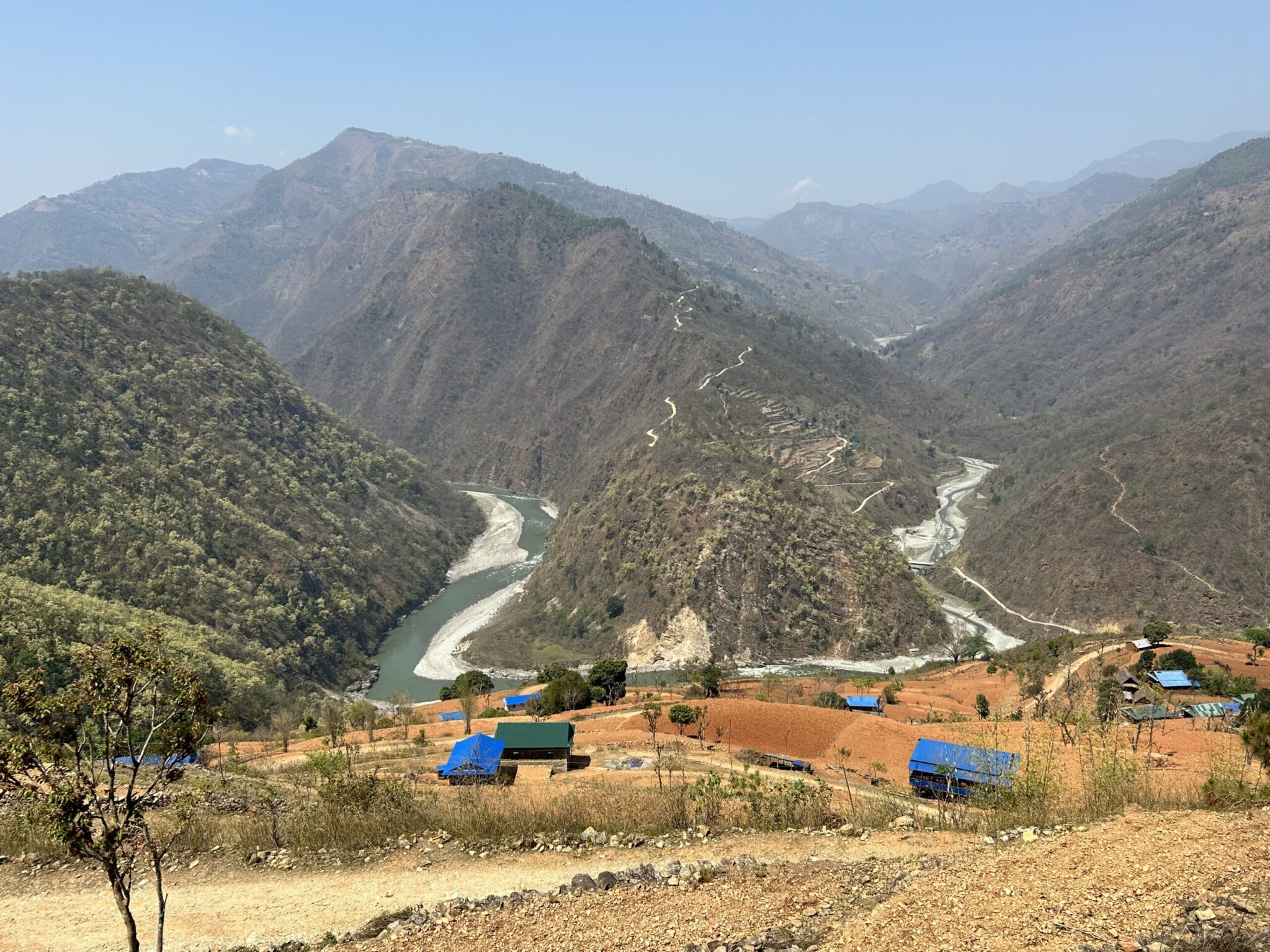 Mountain valley with a winding river, scattered buildings with blue roofs, and dry vegetation in the foreground, surrounded by rugged hills