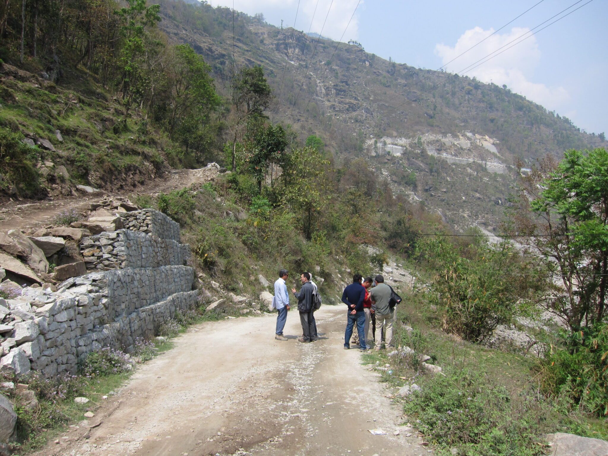 Group of people walking along a dirt mountain road with stone retaining walls, surrounded by vegetation and steep hillsides