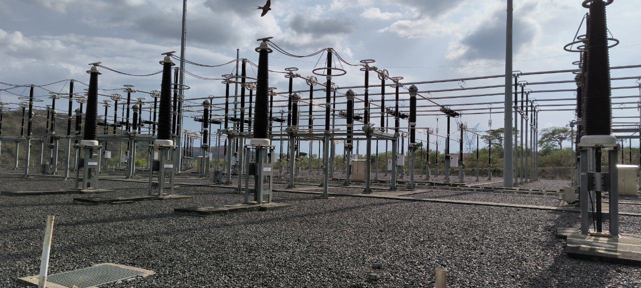 High-voltage electrical substation with multiple circuit breakers and insulators installed on gravel ground, under a cloudy sky