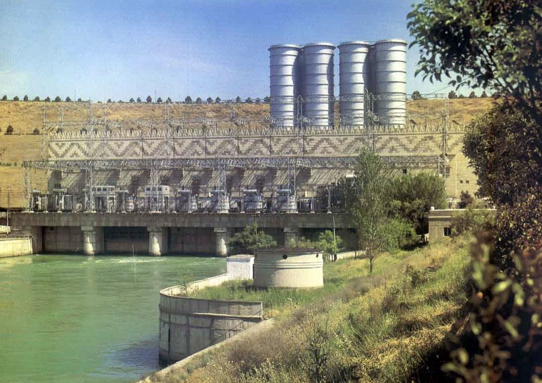 Frontal view of a Hydropower Plant showing the powerhouse structure, water intakes, and large cylindrical draft tubes along a river.