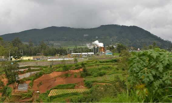 Geothermal power plant emitting steam in a rural agricultural landscape, with terraced fields and forested hills in the background under a cloudy sky