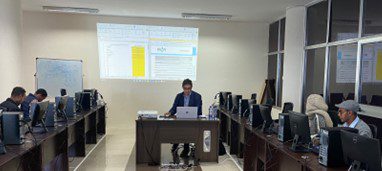 Classroom or training room with people working at desktop computers, while an instructor presents data projected on the wall