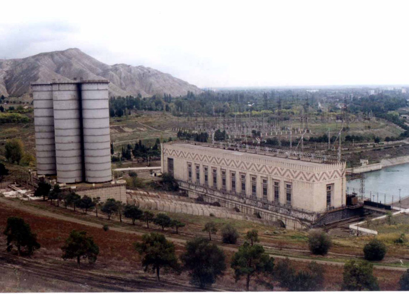 Hydroelectric power plant facility with two large vertical silos and a rectangular turbine hall building, set in a rural landscape near a mountain