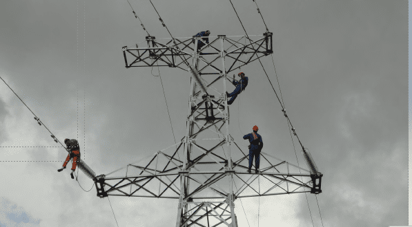 Workers performing maintenance on a high-voltage transmission tower, suspended on power lines against a cloudy sky