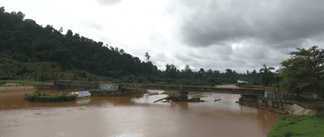 View of a muddy river with a small dam or water control structure, surrounded by tropical vegetation under a cloudy sky
