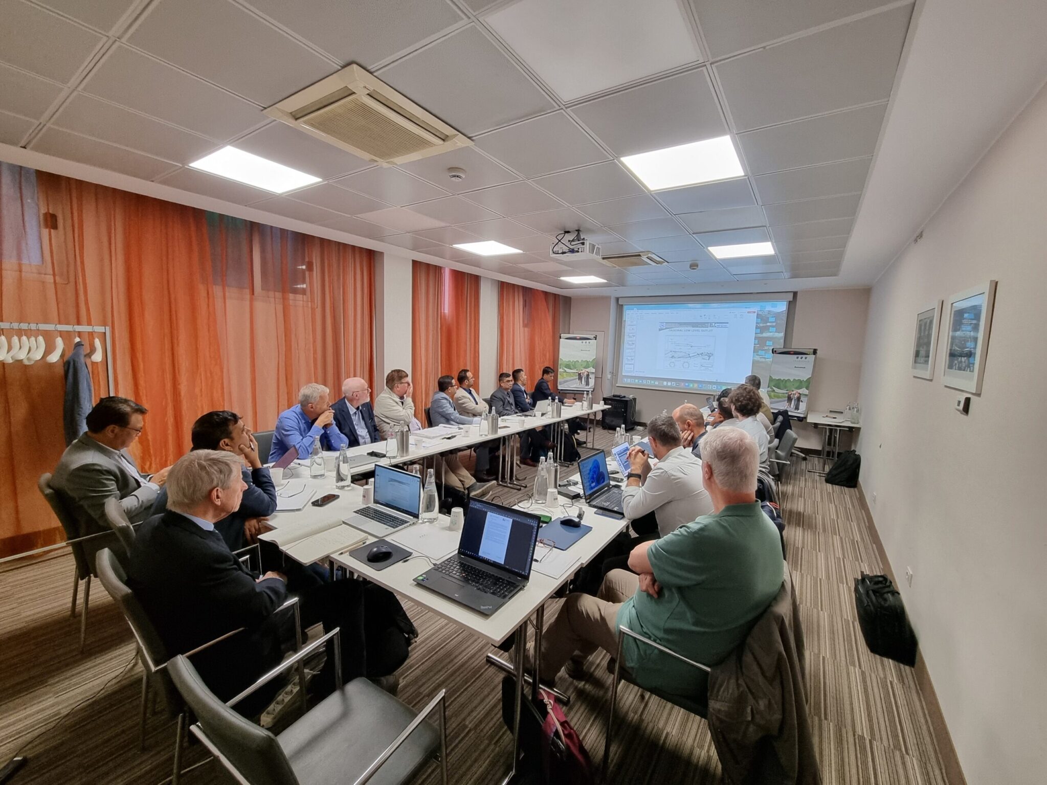 Business meeting or technical workshop with participants seated around a U-shaped table, laptops open, and a presentation projected on the screen