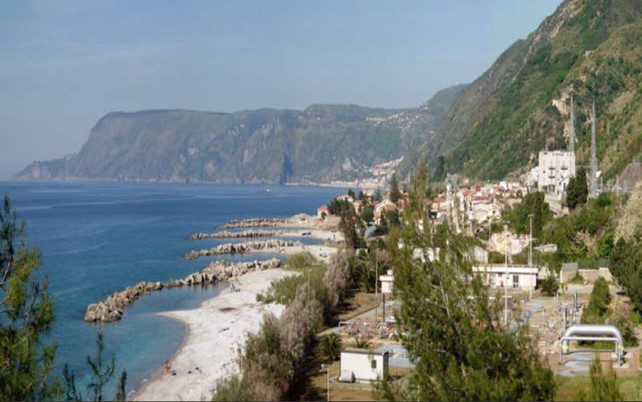 Coastal landscape with a seaside village, sandy beach, breakwaters, and steep green hills descending to the blue sea