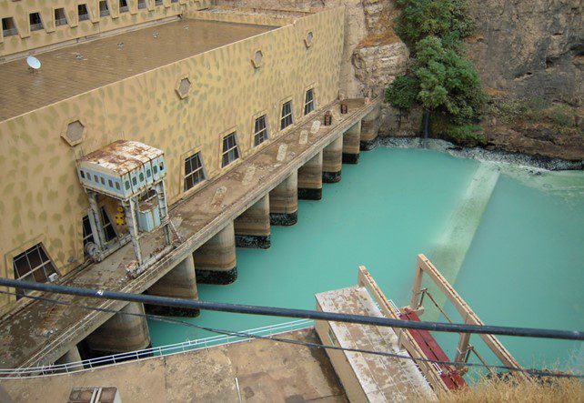 View of a hydroelectric power station with multiple intake gates and turquoise water, located at the base of a rocky cliff