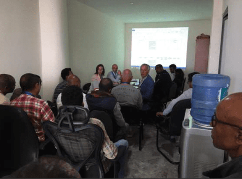 Small group of people attending a presentation in a compact meeting room, with a projected slide on the wall and a water dispenser visible in the foreground