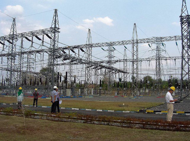 "People inspecting or maintaining a high-voltage electrical substation, with lattice steel structures, power lines, and insulators visible in the background