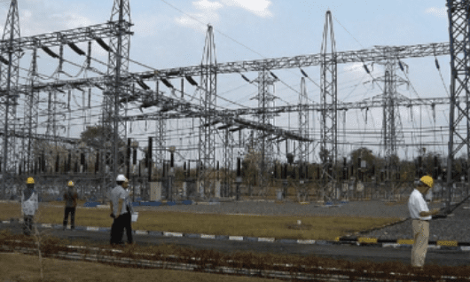 Workers at an electrical substation with high-voltage transmission lines in the background, under a partly cloudy sky.