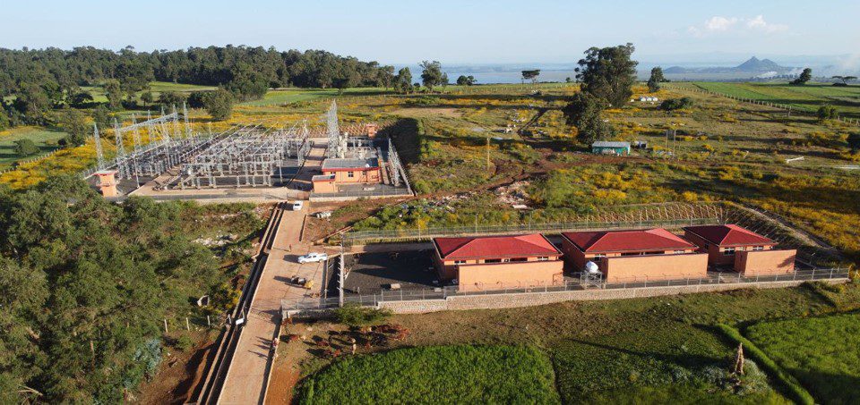 Aerial view of an electrical substation with red-roofed buildings and metal structures, surrounded by green agricultural fields and trees