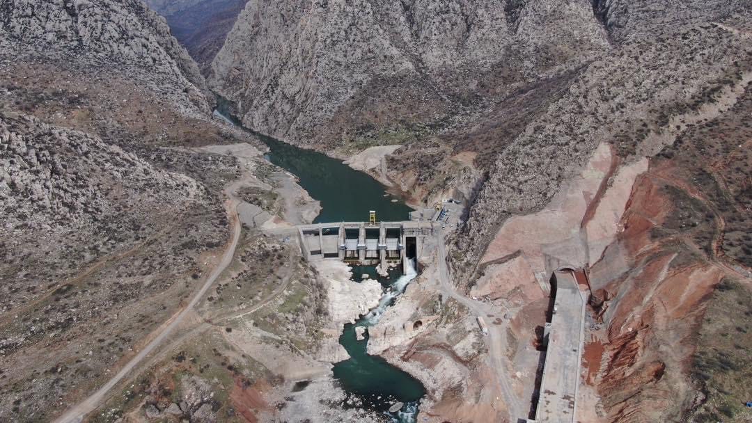 Aerial view of a dam built between steep rocky mountains, with a river flowing through the valley and visible construction works nearby