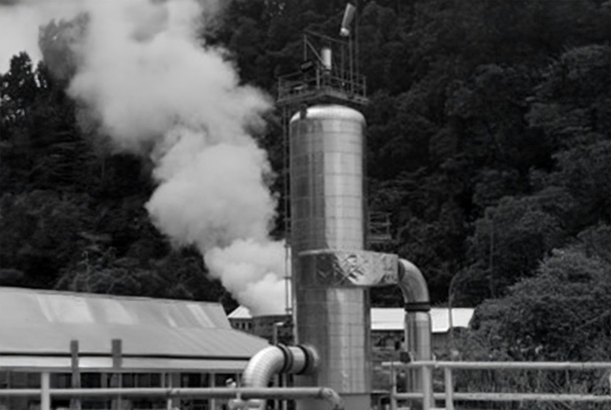 Industrial facility with a vertical metal pipe emitting steam, set against a forested background