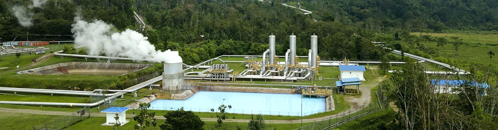 Aerial view of a geothermal power plant with steam rising from wells, cooling towers, water basins, and surrounding green landscape