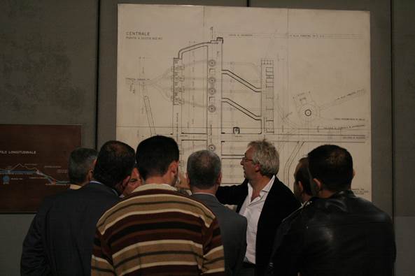 Group of people observing and discussing a large technical architectural drawing displayed on a wall, with a presenter pointing at the plan