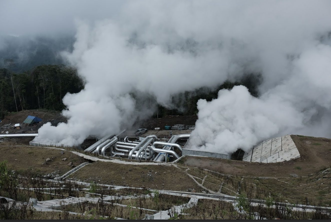 Geothermal power facility set in a mountainous, forested landscape, with visible steam rising from pipelines in the foreground.